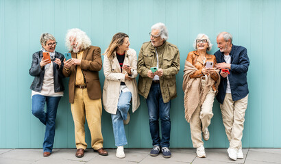 Group of senior people using technology devices together standing on a blue wall - Happy older...
