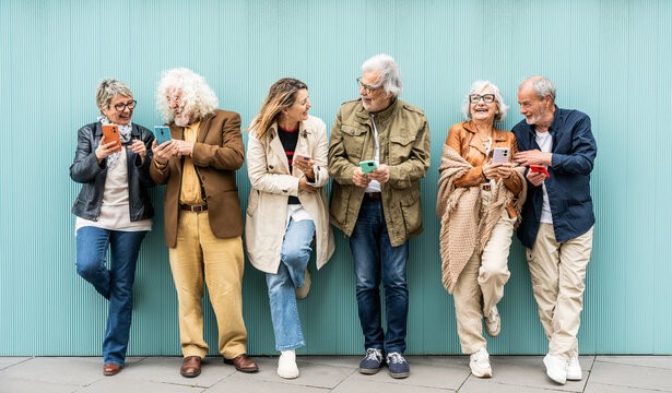 Group of senior people using technology devices together standing on a blue wall - Happy older friends having fun watching funny video on smartphone - Tech and modern elderly concept