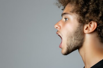 Fototapeta premium Side profile of young mixed race man with curly hair expressing shock and surprise with open mouth against gray background, studio portrait.