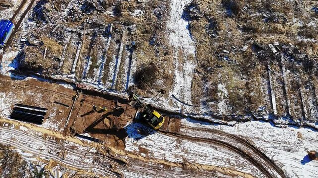 A yellow excavator carves into snow-dusted earth at a construction site in Saliena, Pinki village, revealing muddy paths and exposed ground amid winter conditions.