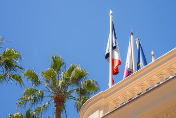 A building with intricate architecture displays French and EU flags, set against a bright blue sky...