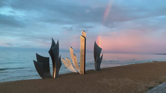 Wide static shot of Les Braves WWII memorial by Anilore Banon on Omaha Beach, Normandy, France, at sunset with a rainbow over the calm sea, pastel sky and sand foreground, no people, camera static