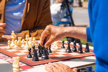A casual chess game between two individuals, one in a blue sweater and the other in a brown jacket, with sunlight and outdoor seating in the background.