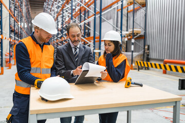 Manager showing documents to warehouse workers standing at table wearing safety vests and helmets