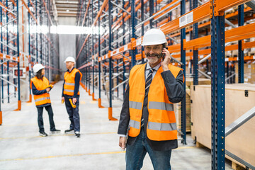 Warehouse manager wearing safety vest and helmet using smartphone and holding tablet, coordinating...