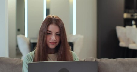 Redhead woman sitting on couch in living room in new apartment and using laptop notebook. Woman is working from home, online shopping. Modern technology, internet and people concept. - Powered by Adobe
