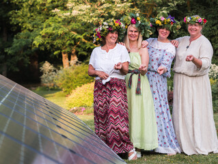 Four women, adorned with homemade wreaths, stand in the garden on a summer solstice evening, with a...