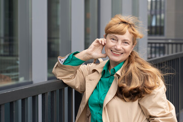 A 60-year-old woman, wrapped in a beige raincoat, walks among tall buildings. Lady smiles and looks at the camera. Concept of beautiful old age and active longevity.