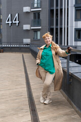 A 60-year-old woman in a beige raincoat walks among tall buildings. Full length view. Urban style. Concept of beautiful old age and active longevity.