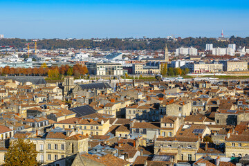 Panoramic elevated view of central and northeastern Bordeaux, showing dense old city rooftops, with a river, bridge, and hills with buildings in the background under a blue sky. Bordeaux, France.
