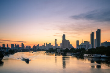 Sunrise view of the Chao Phraya River with skyline silhouettes modern high-rises, as a boat moves through golden reflections.