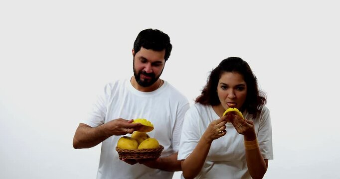 Indian young couple eating mango while standing isolated against white background, holding, promoting fresh seasonal aam or Alphonso happily, enjoying king of fruits with joyful expressions smiles