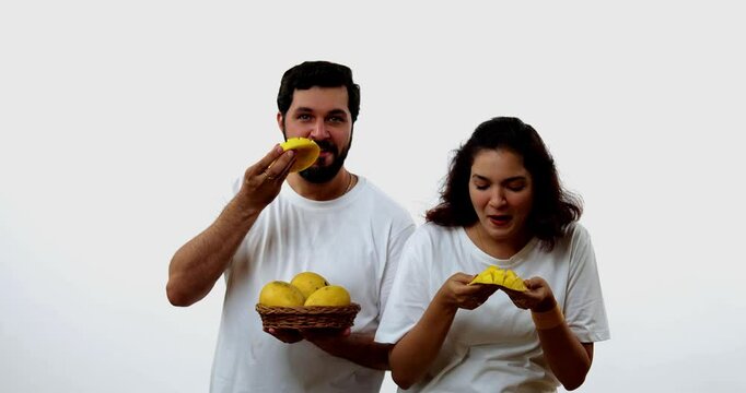 Indian young couple eating mango while standing isolated against white background, holding, promoting fresh seasonal aam or Alphonso happily, enjoying king of fruits with joyful expressions smiles
