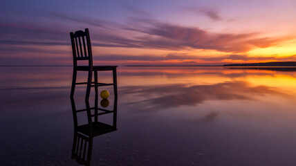 A surreal photograph of a black wooden chair standing in shallow water at sunset.
