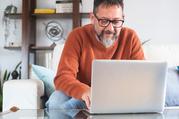 Mature man working from home on laptop, wearing orange sweater. Middle-aged freelancer using technology in domestic workspace. Various expressions from happy to thoughtful. Remote work, focus and life