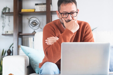 Mature man working from home on laptop, wearing orange sweater. Middle-aged freelancer using...