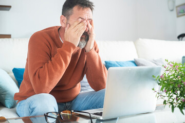Fototapeta premium Mature man working from home on laptop, wearing orange sweater. Middle-aged freelancer using technology in domestic workspace. Various expressions from happy to thoughtful. Remote work, focus and life