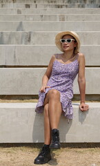 Stylish Asian woman wearing a purple floral dress, black boots and a straw hat sits on concrete steps