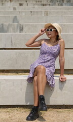 Stylish Asian woman wearing a purple floral dress, black boots and a straw hat sits on concrete steps