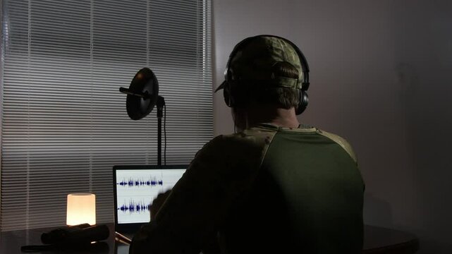 A military officer in camouflage uniform conducts an audition in an isolated environment. Professional audio equipment is used - headphones, microphone and laptop. Concept - intelligence, espionage, 