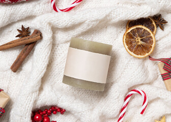 Green pillar candle with blank label on white knitted sweater near Christmas decor, winter mockup