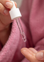Young woman in pink bathrobe dropping essential oil on a finger and holding a pipette closeup