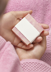 Closeup of young woman in pink bathrobe holding soap bar with blank label, mockup