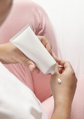 Woman in pink pajama squeezing cream on hand and holding a cosmetic tube, closeup