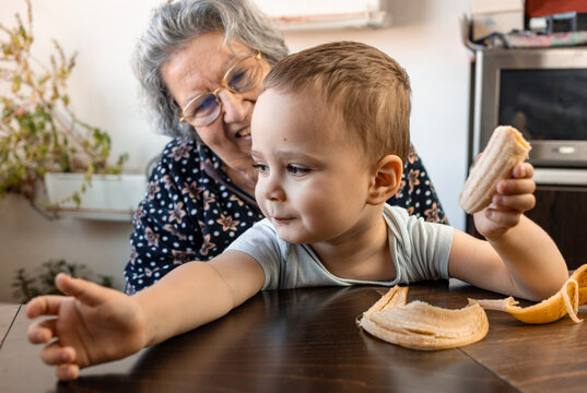 2,5 YO Jewish child holding banana sitting in his 80 year old grandmother on the table, smiling..