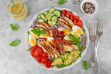 Healthy cobb salad with roasted chicken breast, chopped avocado, tomatoes, bacon, cucumber, hard boiled egg and green leaves on a plate. Top view.
