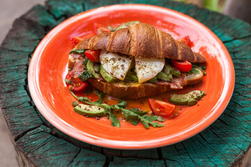 Homemade croissant sandwich with avocado, poached egg, bacon, arugula, and cherry tomatoes. A stylish brunch with a child enjoying the meal