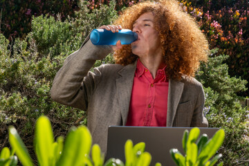 Detail of a Young man with curly hair drinking from a reusable bottle while working on a laptop outdoors, surrounded by greenery. Concept of sustainable remote work and eco-friendly lifestyle.