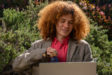 Young man with curly hair working on a laptop outdoors, holding a reusable bottle. Concept of sustainable remote work, eco-friendly lifestyle and digital wellbeing.
