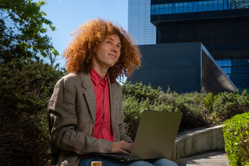 Young man with curly hair working on a laptop outdoors, looking up with a thoughtful expression. Concept of sustainable remote work, creativity, eco-friendly business and future vision.