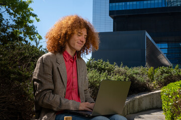 Young man with curly hair working on a laptop outdoors, surrounded by greenery and modern architecture. Concept of sustainable remote work, urban wellbeing and eco-friendly business lifestyle.