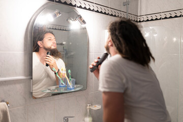 Young man shaving beard with electric razor in bathroom mirror