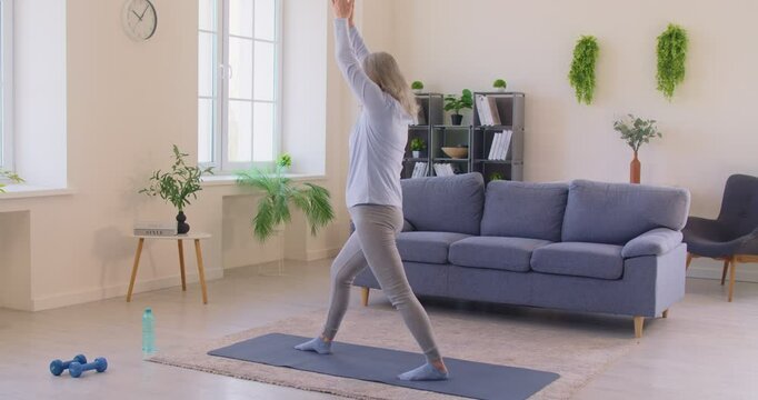 Old woman doing stretching exercises at home, keeping arms raised over head, standing on fitness mat, doing morning sports, training indoor at home. Senior people and healthy lifestyle concept