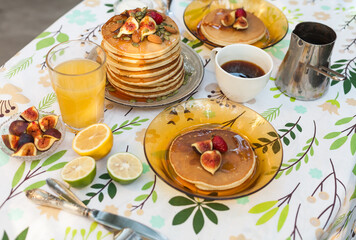 Stack of pancakes with fresh fig, strawberries, maple syrup, and crunchy toppings. A warm, comforting outdoor breakfast with orange juice and coffee