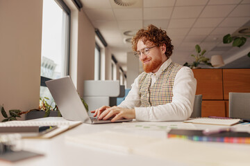 A young man with vibrant red hair is using his sleek laptop in a bright, modern office, showcasing remote work culture