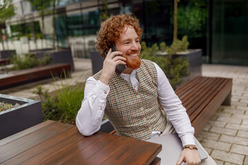 A stylish man with a groomed beard enjoys a lively phone call outdoors, radiating confidence and...