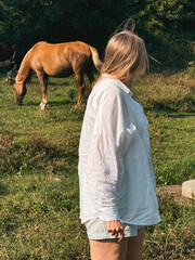 Blonde woman in white shirt looking at horse grazing on grass