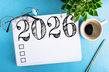 New year resolutions 2026 on desk. 2026 goals list with notebook, coffee cup, plant on blue table. Resolutions, plan, goals, action, checklist, idea concept. New Year 2026 resolutions. Copy space