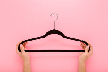 Young adult woman hands holding and showing dark black velvet empty hanger without clothes on light pink table background. Pastel color. Closeup. Point of view shot. Top down view.