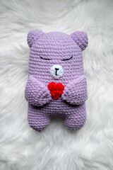 A handmade purple teddy bear with its eyes closed, gently holding a small heart. The bear is placed on a soft, fluffy white background, creating a cozy and tender atmosphere. 