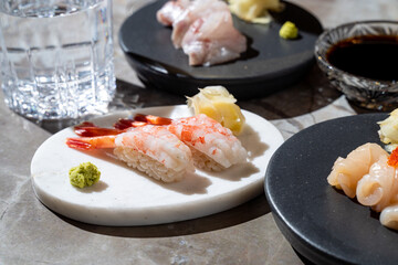 A beautifully arranged sushi platter showcases delicate shrimp sushi nestled on a white plate. Nearby, a glass of water and bowls of soy sauce and wasabi complete the culinary experience