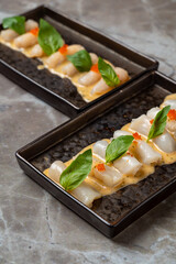 Two sleek black plates display a beautifully arranged seafood dish, featuring scallops topped with a creamy sauce, bright garnishes of basil, and colorful roe, set against a textured stone surface