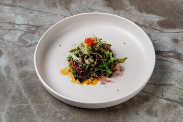 Intricately arranged salad featuring a variety of greens, topped with colorful garnishes and a light dressing. The dish is presented on a sleek, simple plate, embodying a contemporary culinary style