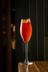 A vibrant cocktail adorned with a twist of orange sits elegantly on a polished wooden surface, reflecting the warm ambiance of a chic bar. The rich colors invite indulgence and leisure