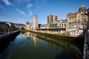 Obraz premium Cityscape of Bilbao shimmering in the Nervion river, featuring stunning reflections under a bright blue sky filled with clouds