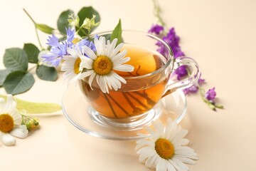 Aromatic herbal tea in glass cup and different flowers on beige background, closeup
